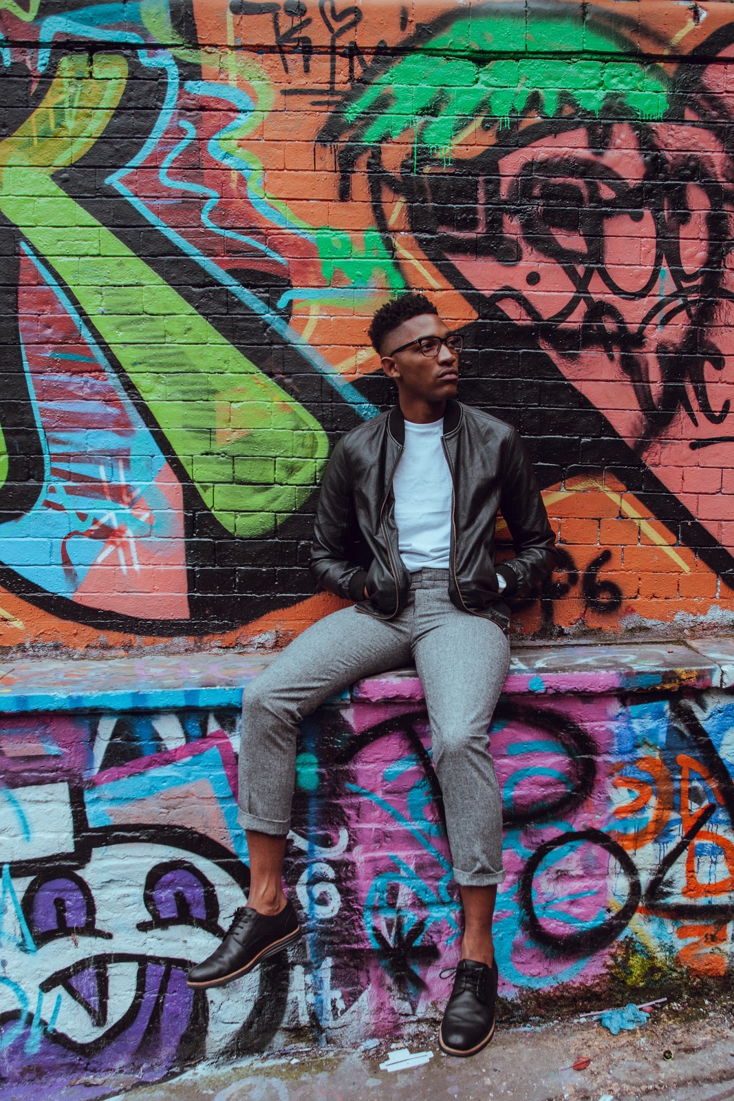 A young man casually sits on a ledge with graffiti behind him. He is wearing a leather bomber jacket, white shirt, grey pants, and black oxford shoes.